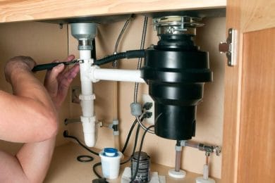 Plumber repairing a garbage disposal beneath a kitchen sink in Brighton